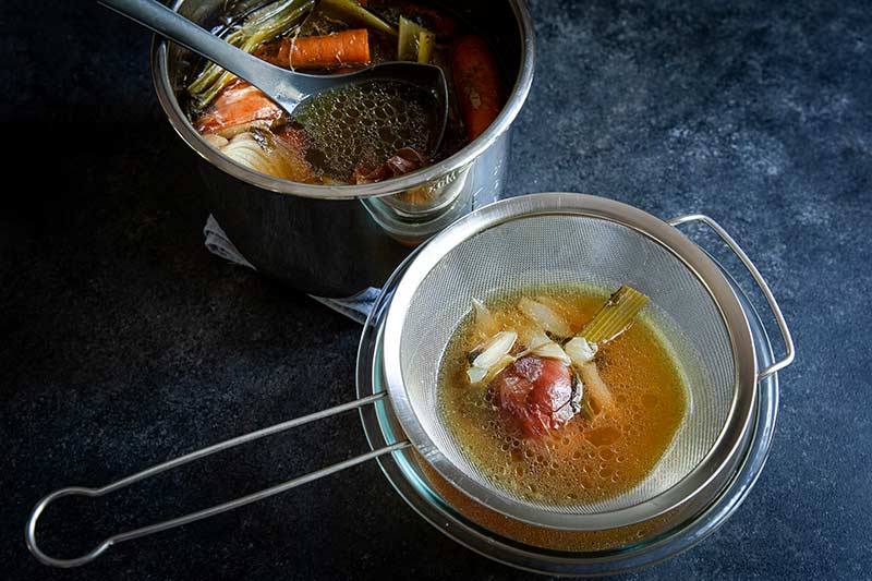 A fine mesh strainer set over a bowl with homemade chicken stock ladled in