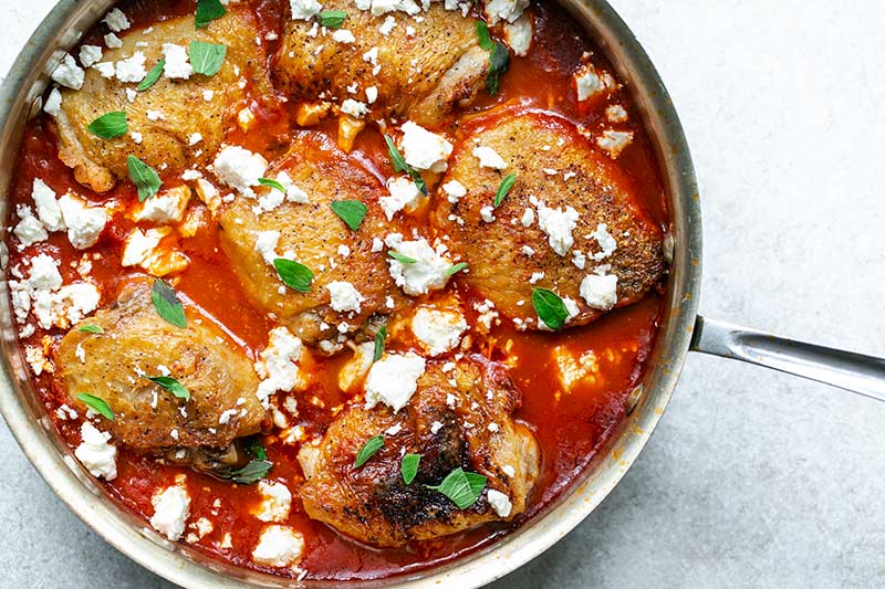 A closeup of chicken thighs in skillet with tomato sauce, topped with feta and oregano