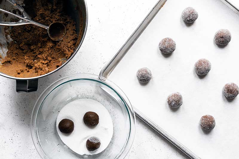 A mixer with cookie dough, a bowl of sugar for tossing raw cookies, and a sheet pan with unbaked molasses cookies