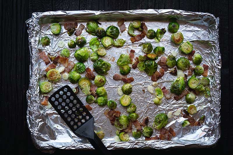 A foil-lined sheet pan topped with partially roasted brussels sprouts, bacon, olive oil, sliced garlic, salt and pepper.