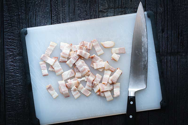 Cutting bacon into strips on a cutting board with a chef’s knife