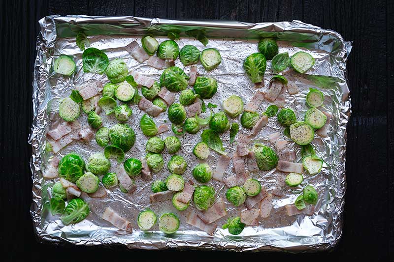 A foil-lined sheet pan topped with raw brussels sprouts, bacon, olive oil, salt and pepper.