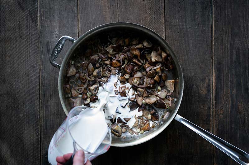A hand pouring cream into the mushrooms and shallots.