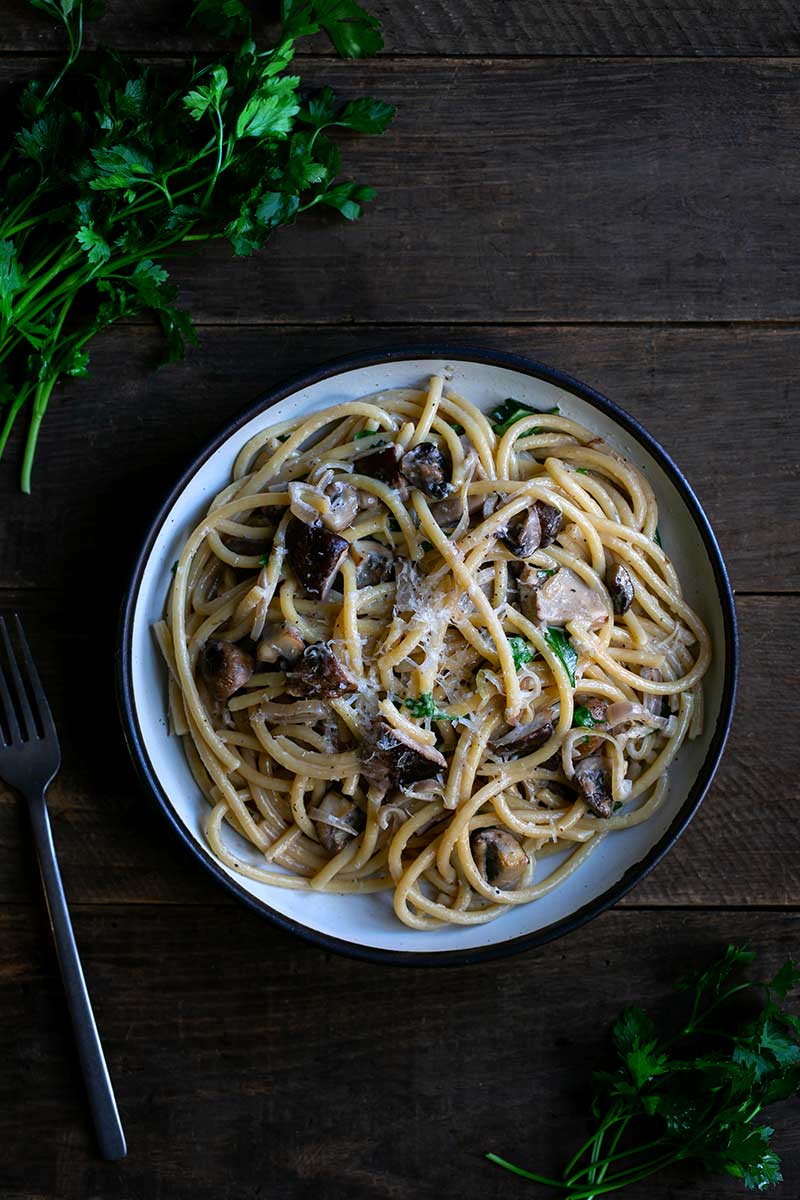 Creamy mushroom pasta recipe on a plate next to a fork and bunches of fresh parsley.