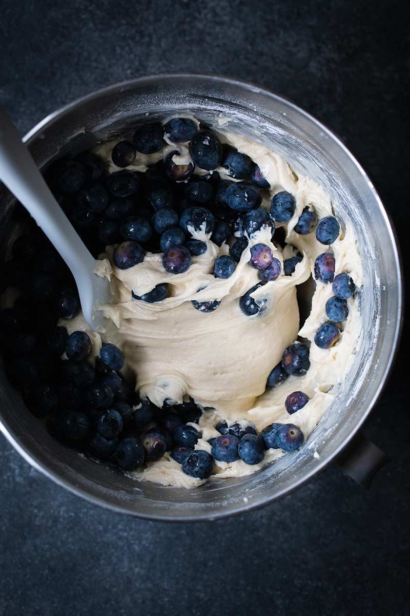 Cake batter in a stand mixer with blueberries mixed in using a spatula